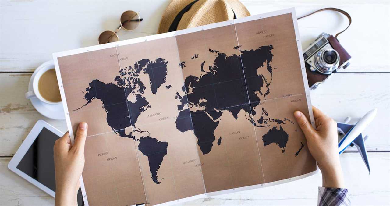 map of the world in a person's hands with a toy plane, camera, coffee and tablet surrounding the brown and black map