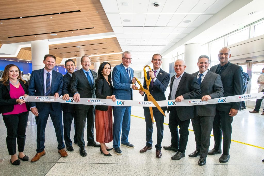 Airline staff stand in a row, smiling. Man in the centre cuts ribbon with large scissors.