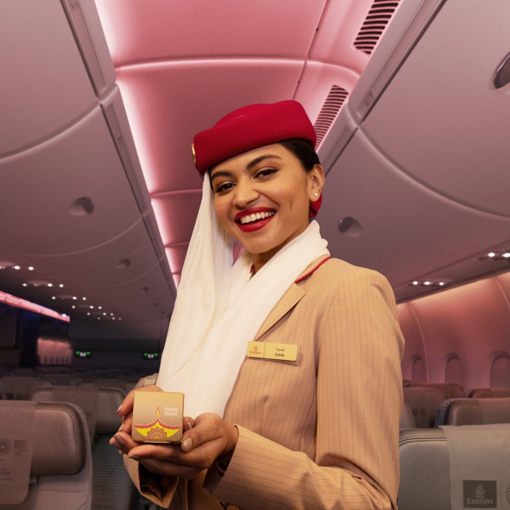 Emirates flight attendant with red lipstick smiles in cabin, holding small box in hands.