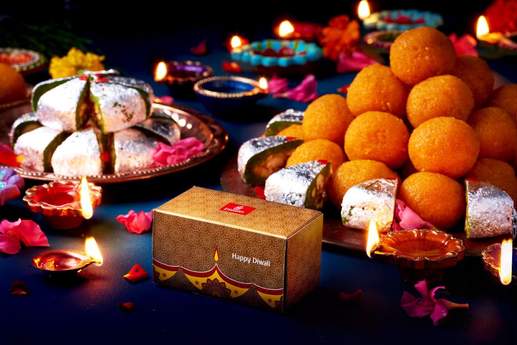 A selection of Diwali sweets arranged on a dark table cloth, surrounded by diya lights.