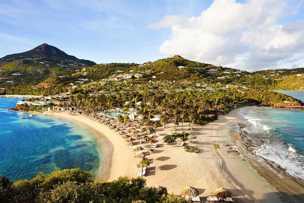 aerial view of beach resort with lush green trees, chairs along the water, and mountains in the distance.