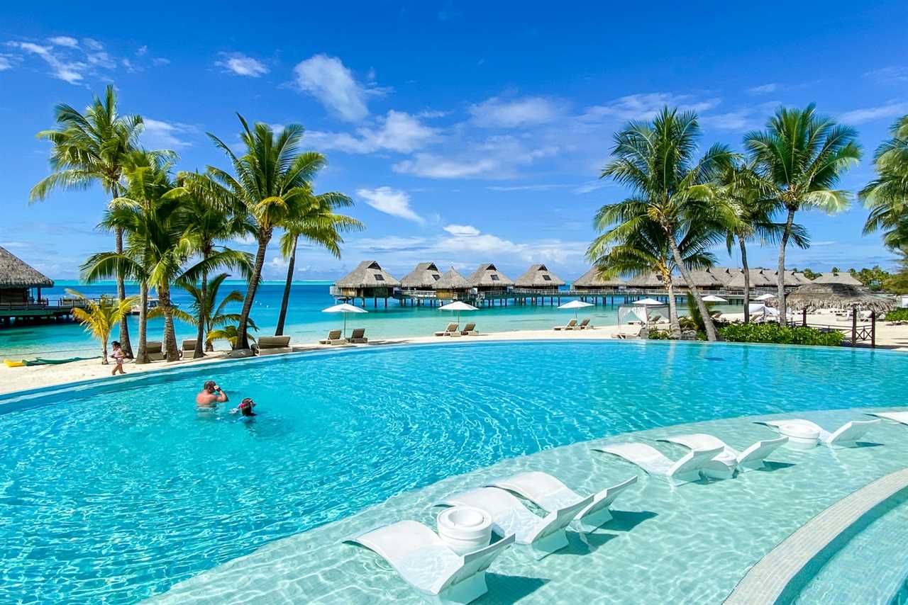 a swimming pool and lounge chairs with overwater bungalows visible in the background, showing parts of a luxury hotel on an island