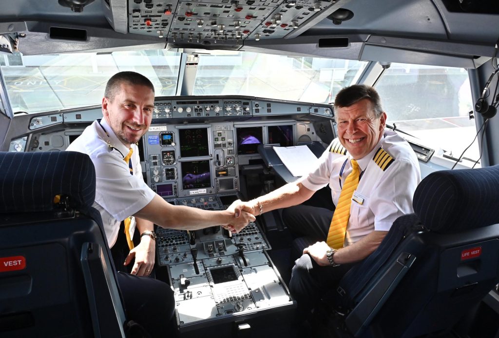 Two male pilots in cockpit look into camera, shaking hands.