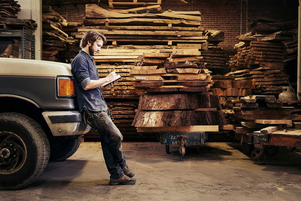 a person leans against a pickup truck, making notes on a notepad while surrounded by piles of wood in a carpentry shop