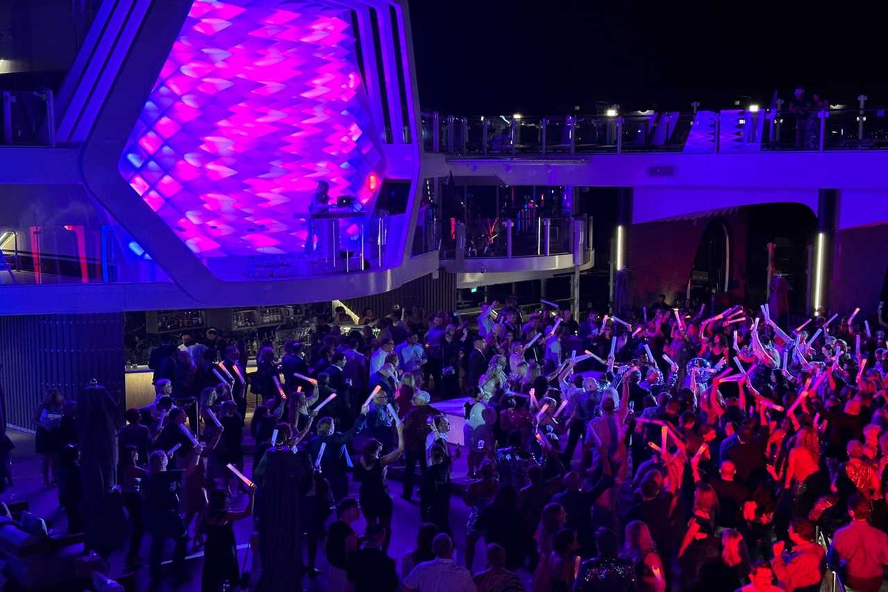 A dancing crowd gathered in front of a DJ booth for a party on the pool deck of a cruise ship at night