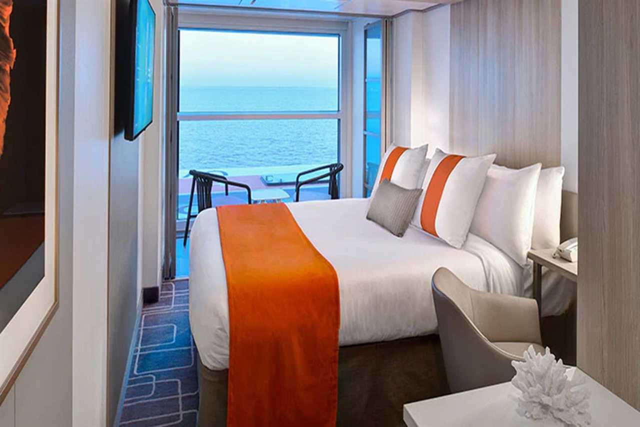 A queen bed in a small cruise ship cabin. It's adorned with several throw pillows and an orange bed runner.