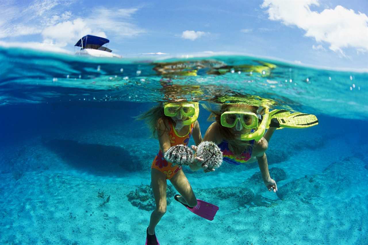 Partial underwater photo of two girls in neon yellow snorkels and goggles holding a sea creature while a boat bobs nearby